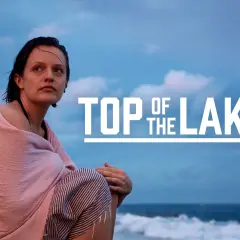 Kobieta owinięta w różowy ręcznik siedzi na plaży, patrząc w dal. Na niebie widnieje napis "TOP OF THE LAKE".