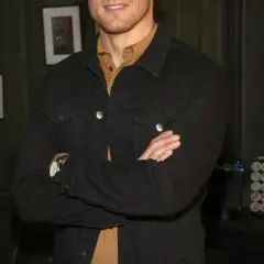 Sam Heughan w czarnej kurtce dżinsowej, z rękami skrzyżowanymi na piersi. W tle zdjęcia i ekspres do kawy.