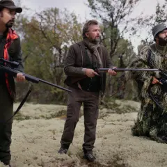 Trzech myśliwych w kamuflażu i odzieży myśliwskiej, z bronią w rękach, przemierza odludzie.