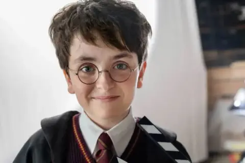 Chłopiec w okularach i szacie Hogwartu, trzymający klaps filmowy. Wygląda jak nowy Harry Potter, gotowy na przygodę.