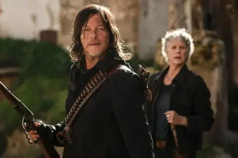 Daryl i Carol, bohaterowie seriali o zombie, przygotowują się do walki. Daryl trzyma strzelbę, a Carol stoi za nim.