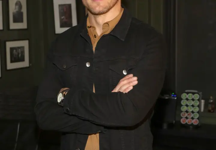 Sam Heughan w czarnej kurtce dżinsowej, z rękami skrzyżowanymi na piersi. W tle zdjęcia i ekspres do kawy.