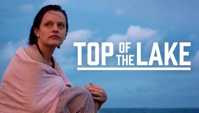 Kobieta owinięta w różowy ręcznik siedzi na plaży, patrząc w dal. Na niebie widnieje napis "TOP OF THE LAKE".