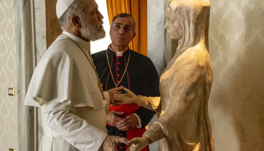 Papież w białej sutannie i kardynał w czerni rozmawiają przy kamiennej rzeźbie. Scena z serialu "nowy papież serial".