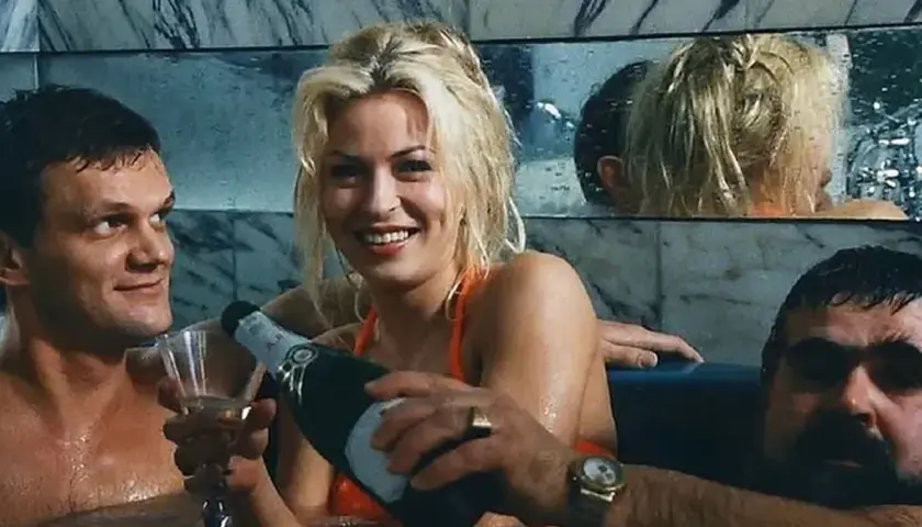 Wspólnik kilera z komedii Machulskiego, uśmiechnięta blondynka nalewa szampana, obok mężczyzna z kieliszkiem.