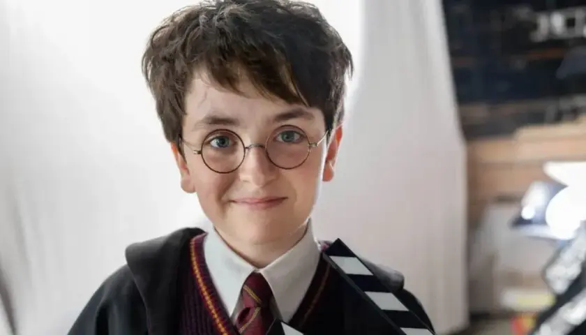 Chłopiec w okularach i szacie Hogwartu, trzymający klaps filmowy. Wygląda jak nowy Harry Potter, gotowy na przygodę.
