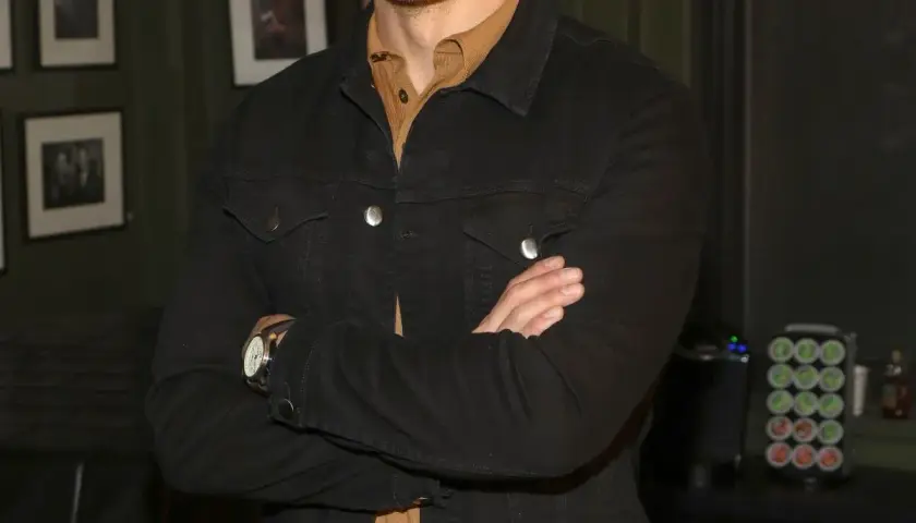 Sam Heughan w czarnej kurtce dżinsowej, z rękami skrzyżowanymi na piersi. W tle zdjęcia i ekspres do kawy.