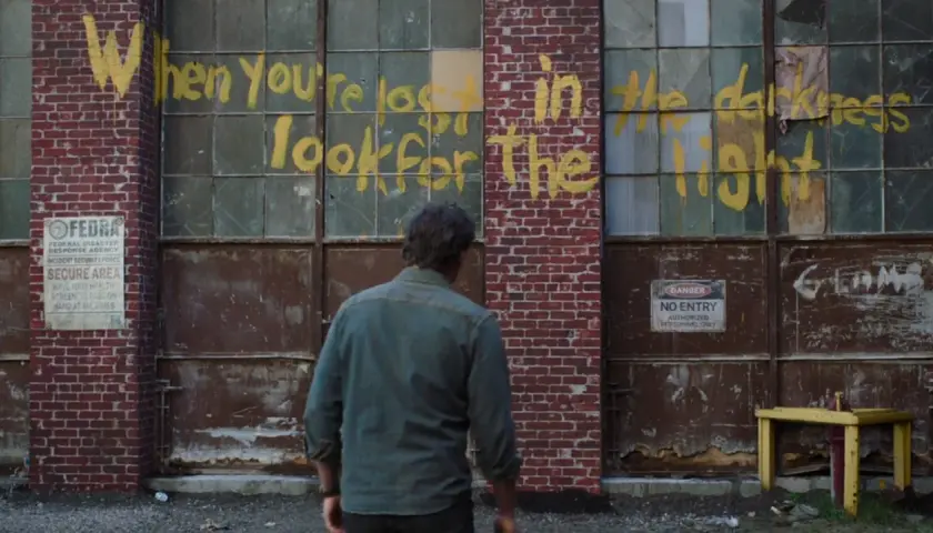 Joel patrzy na napis "When you're lost in the darkness, look for the light" na ścianie budynku, nawiązanie do świetlików z The Last of Us.