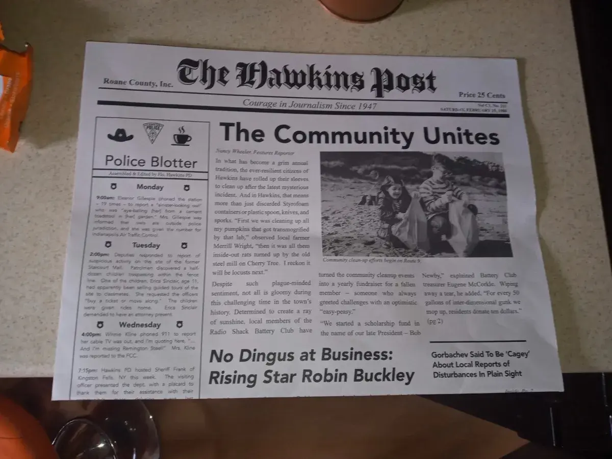 Gazeta "The Hawkins Post" z artykułem "The Community Unites" i zdjęciem dzieci sprzątających plażę.