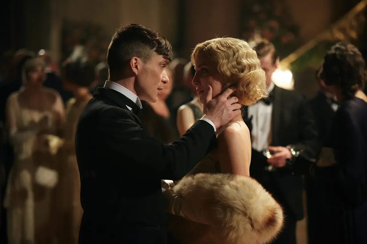 Tommy Shelby czule dotyka policzka Grace, jednej z kluczowych kobiet w świecie Peaky Blinders. W tle widać innych gości balu.