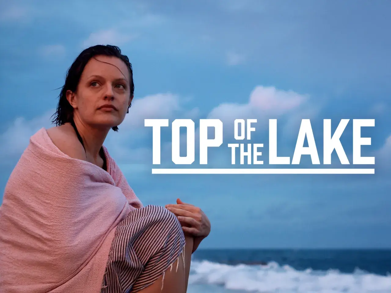 Kobieta owinięta w różowy ręcznik siedzi na plaży, patrząc w dal. Na niebie widnieje napis "TOP OF THE LAKE".