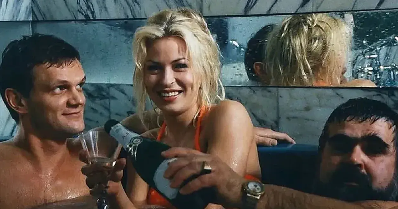 Wspólnik kilera z komedii Machulskiego, uśmiechnięta blondynka nalewa szampana, obok mężczyzna z kieliszkiem.