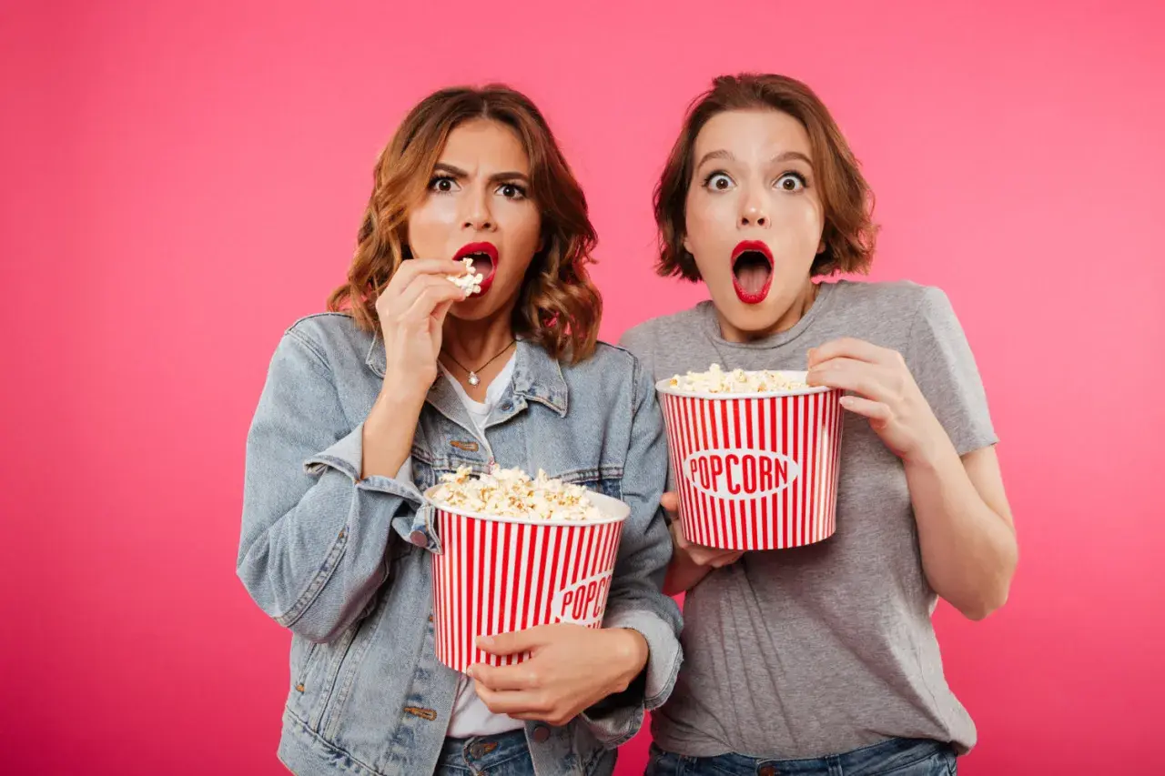 Dwie dziewczyny z wielkimi wiaderkami popcornu, zszokowane i podekscytowane, jakby odkryły, co nowego w kinach.