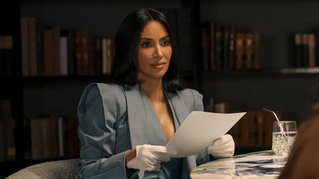 Kim Kardashian w eleganckim kostiumie i białych rękawiczkach czyta dokumenty, jakby przygotowywała się do roli w nowym serialu.