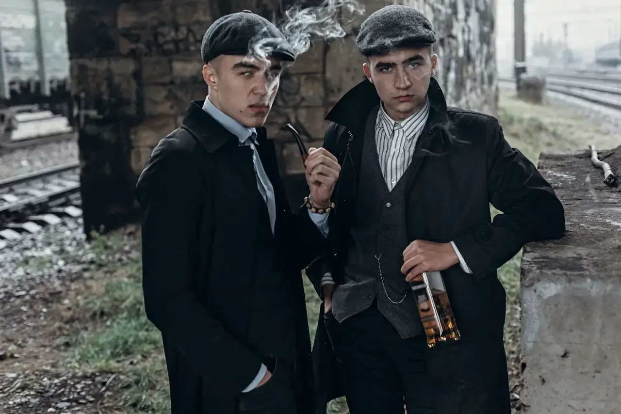 Dwóch młodych mężczyzn w czapkach i płaszczach, jak z seriali o mafii, pali fajkę i trzyma butelkę.