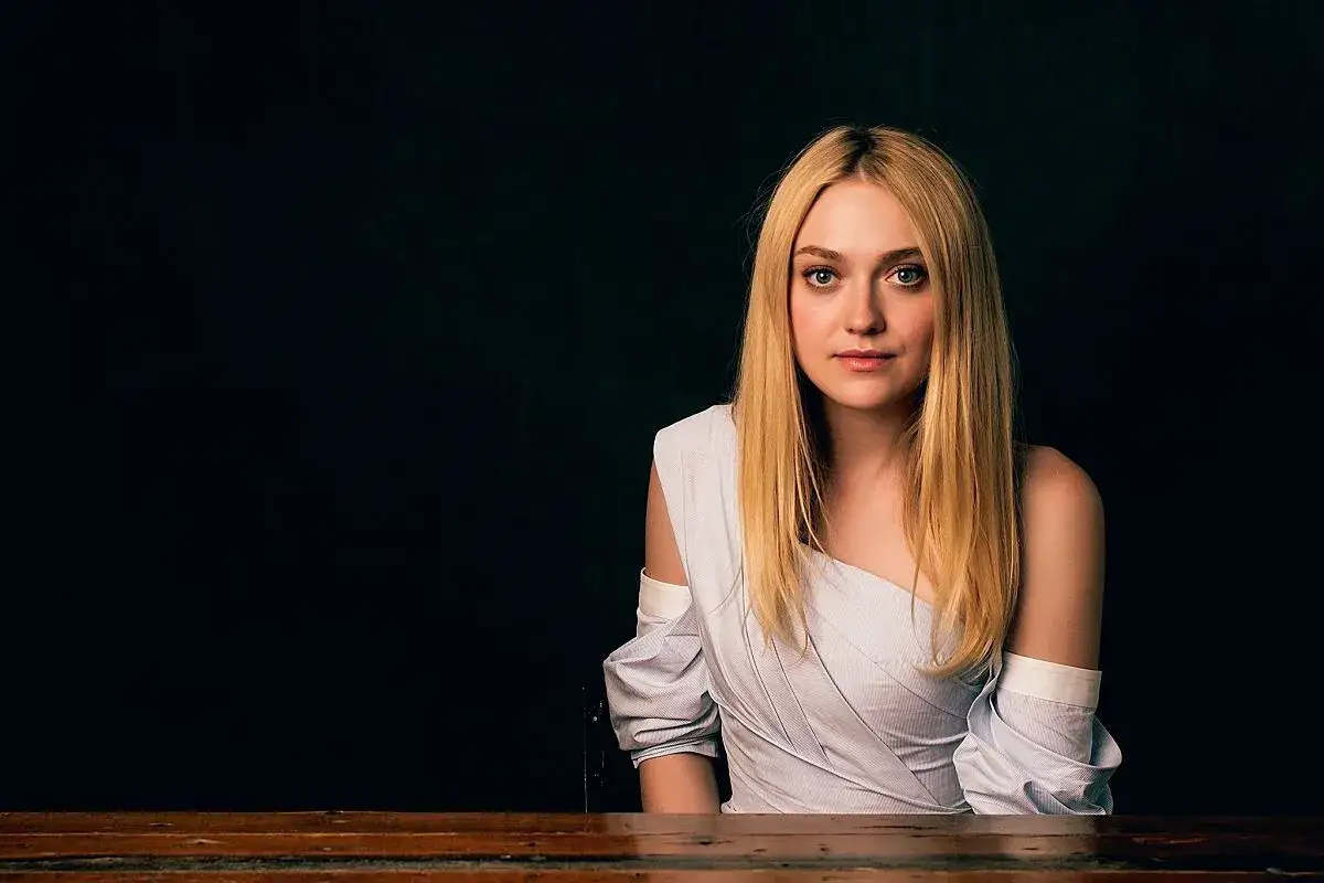 Dakota Fanning, jako dziecko, z blond włosami i niebieskimi oczami, w białej bluzce z odkrytymi ramionami, siedzi przy drewnianym stole.