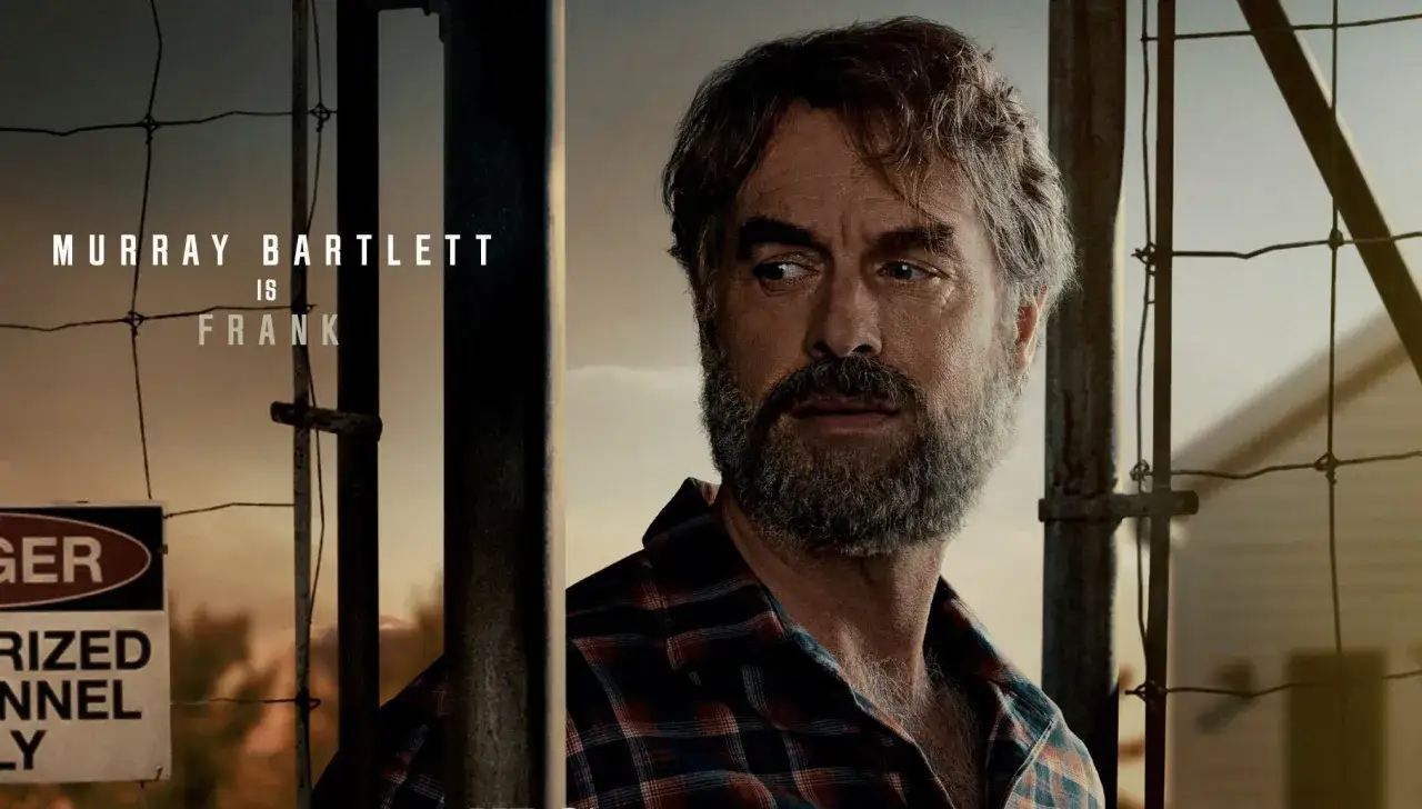 Murray Bartlett jako Frank z The Last of Us. Mężczyzna z siwiejącą brodą i w kratę, za ogrodzeniem.