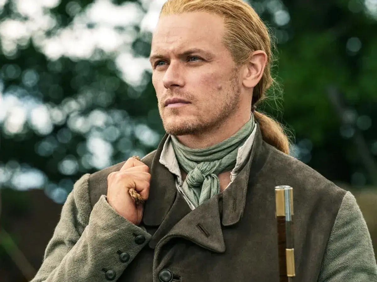 Sam Heughan w stroju z epoki, z blond włosami spiętymi w kucyk, wygląda jakby myślał o swoich dzieciach.