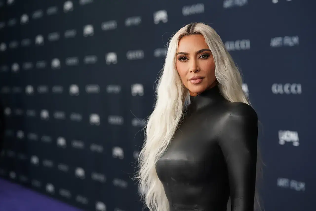 Kim Kardashian w czarnej, obcisłej kreacji na tle z logo Gucci. Jej blond włosy opadają na ramiona, a makijaż podkreśla urodę. Wygląda jak gwiazda z własnego serialu.
