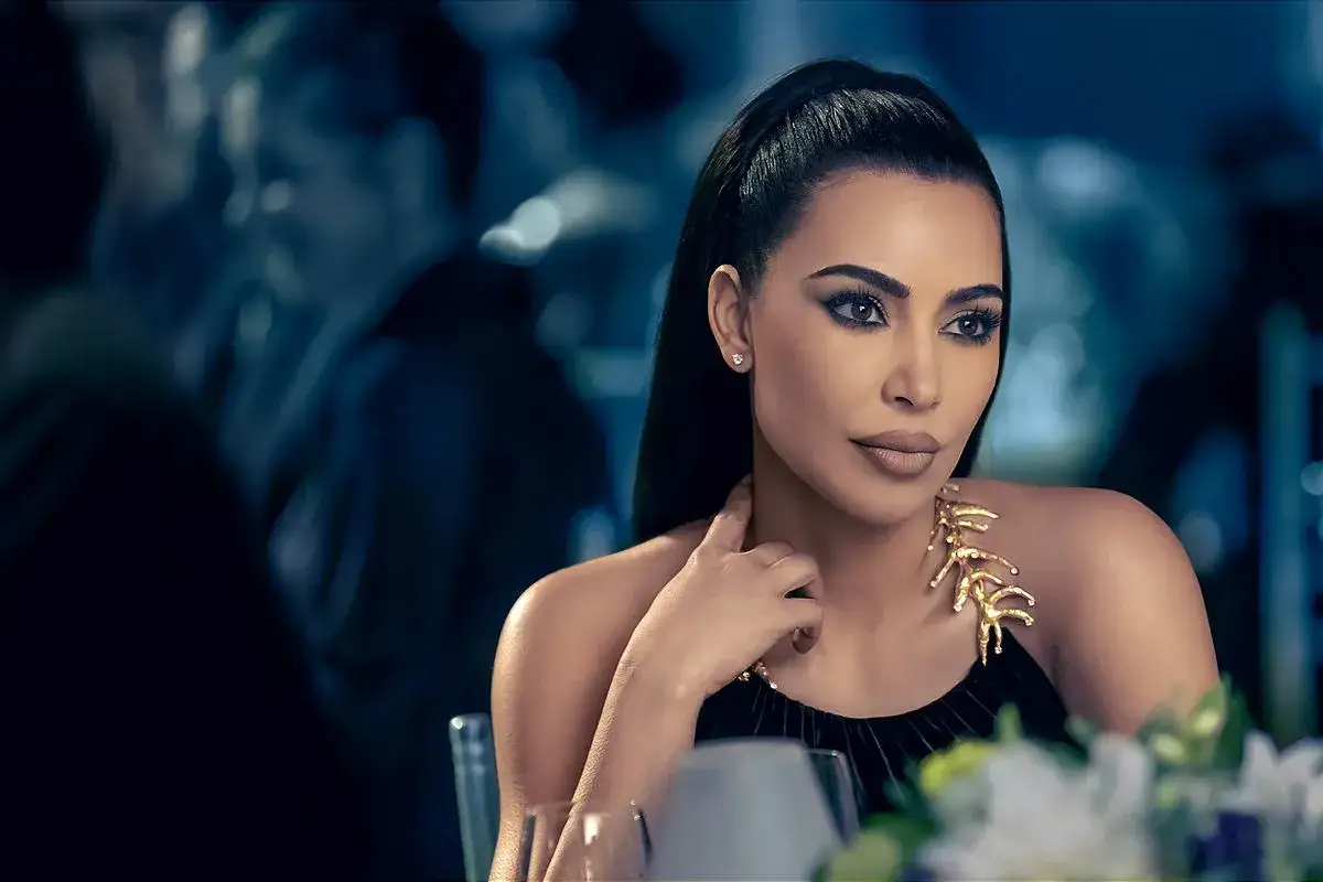 Kim Kardashian w eleganckiej kreacji, z wyrazistym makijażem i złotą biżuterią, wygląda jak gwiazda serialu.