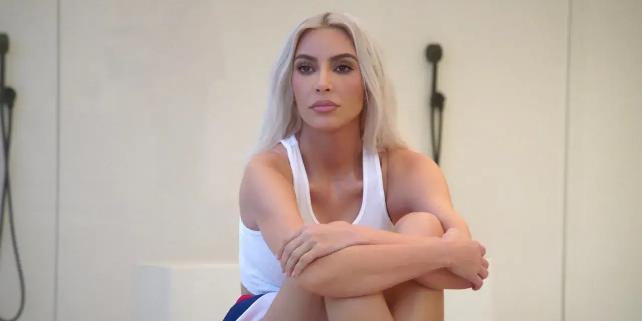 Kim Kardashian w białej koszulce, siedzi ze złożonymi rękami, jakby przygotowywała się do kolejnego odcinka swojego serialu.