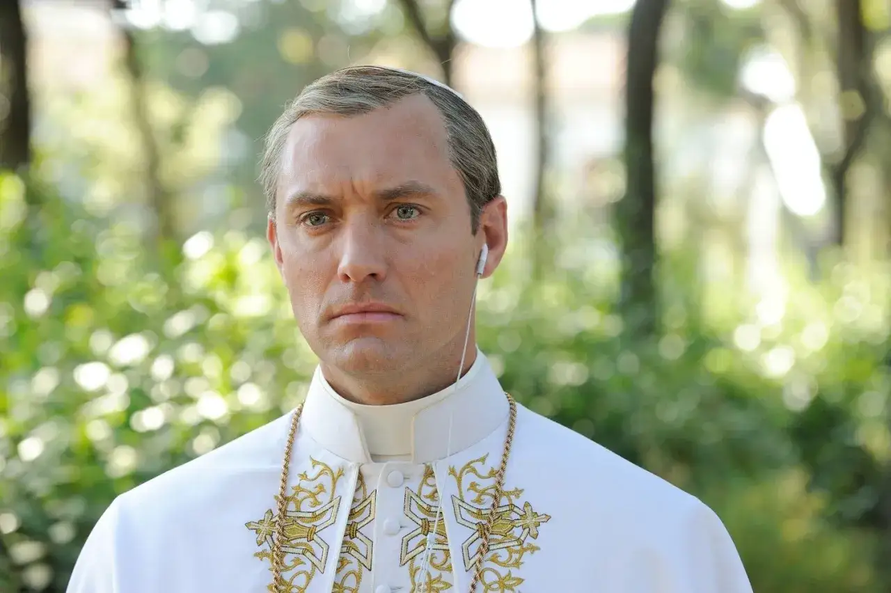 Młody papież, Jude Law, w białej szacie z złotymi zdobieniami, słucha muzyki przez słuchawki. Nowy papież serial.