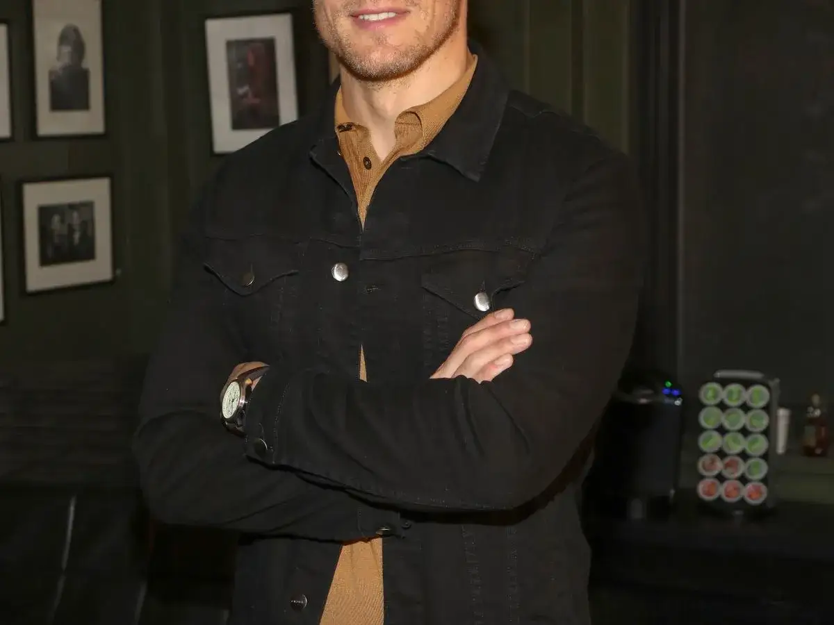 Sam Heughan, w czarnej kurtce i brązowej koszuli, z rękami skrzyżowanymi na piersi. Czyżby myślał o swojej żonie?