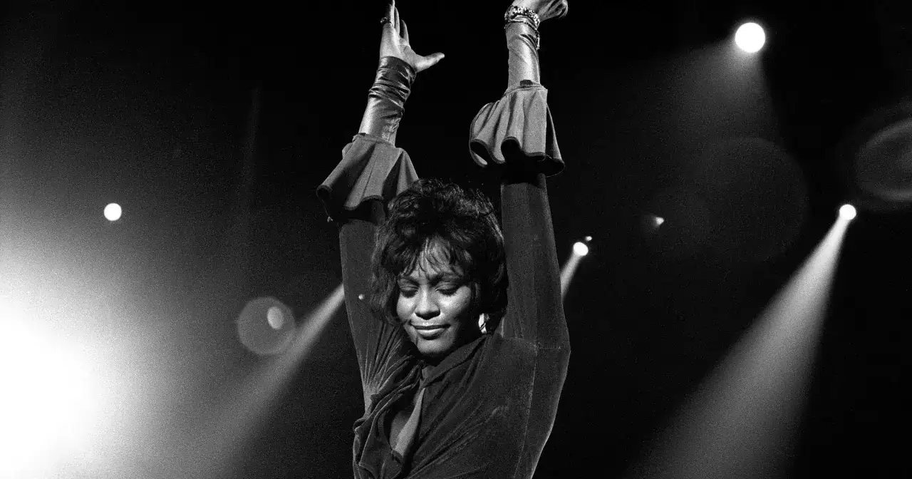 Whitney Houston na scenie, z rękami w górze, w świetle reflektorów. Jej uśmiech sprawia, że zastanawiamy się, czy Whitney Houston żyje.
