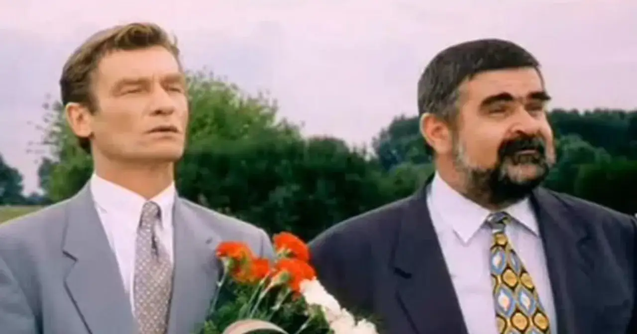 Dwóch mężczyzn w garniturach, jeden z nich to wspólnik kilera z komedii Machulskiego, trzymają bukiet czerwonych kwiatów.